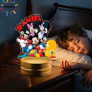 Può includere: Una luce notturna in acrilico personalizzata con un'illustrazione colorata di Topolino, Minnie, Pippo e Paperina. Il nome DANIEL è stampato in rosso nella parte superiore. Il pannello in acrilico poggia su una base rotonda in legno chiaro.