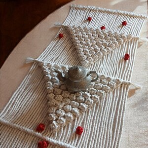 Pode incluir: Um corredor de mesa branco de macramé com um padrão geométrico e pompons vermelhos. Um bule de chá de prata está sobre o corredor de mesa.