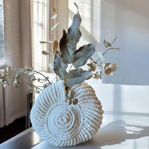 Può includere: Un vaso bianco all'uncinetto con un design circolare, riempito di rami di eucalipto secchi.
