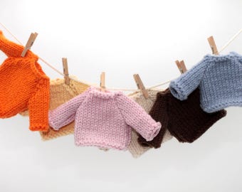 May include: Six miniature knitted sweaters in various colors hanging on a clothesline. The sweaters are orange, yellow, pink, beige, brown, and blue.