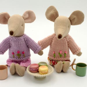 May include: Two plush mouse toys wearing knitted sweaters with embroidered flowers. The mice are sitting on a table with a plate of macarons and two teacups.
