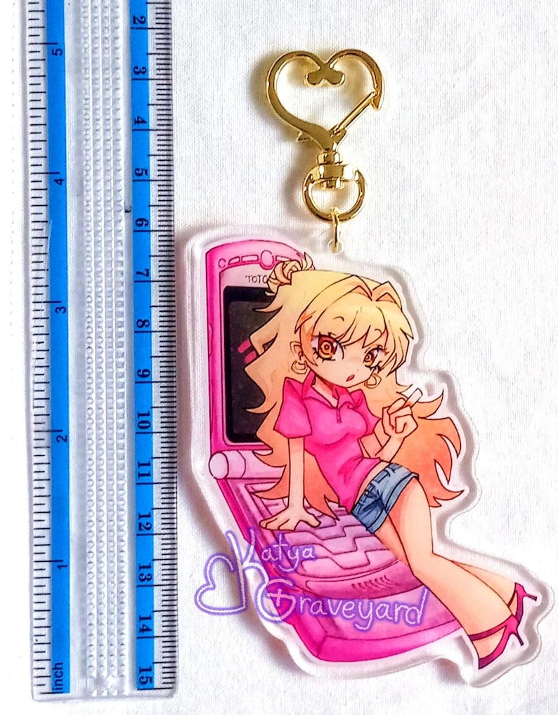 May include: A clear acrylic keychain with a cartoon girl in a pink top and denim shorts, sitting on a pink flip phone. The keychain has a gold-coloured heart-shaped clasp. The text "Katya Graveyard" is visible. The ruler shows inches.