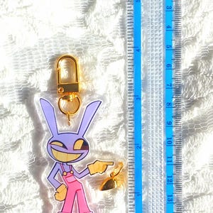May include: A clear acrylic keychain featuring a cartoon rabbit character in purple and pink overalls, with a gold-colored clasp and charm. The keychain is approximately 3 inches long, as measured by the blue ruler in the image.