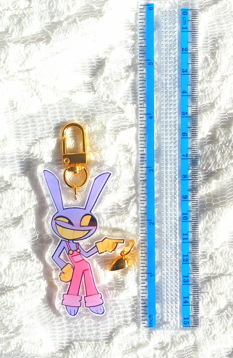 May include: A clear acrylic keychain featuring a cartoon rabbit character in purple and pink overalls, with a gold-colored clasp and charm. The keychain is approximately 3 inches long, as measured by the blue ruler in the image.