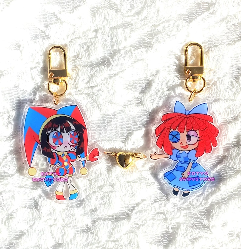 May include: Two acrylic keychains with gold-colored hardware. One keychain features a cartoon jester in red, blue, and yellow. The other keychain depicts a cartoon doll in a blue dress and red hair. A gold heart charm connects the keychains.