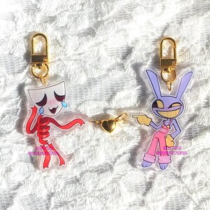 May include: Two acrylic keychains with gold-colored hardware. One features a crying character with a white mask and red body. The other is a purple bunny character in pink overalls. A gold heart charm connects them.