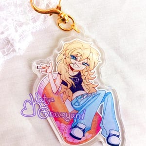 May include: Clear acrylic keyring with a cartoon illustration of a person with blonde hair, blue eyes, and blue jeans. The keyring has a gold heart-shaped clasp and the text "Katya Graveyard".