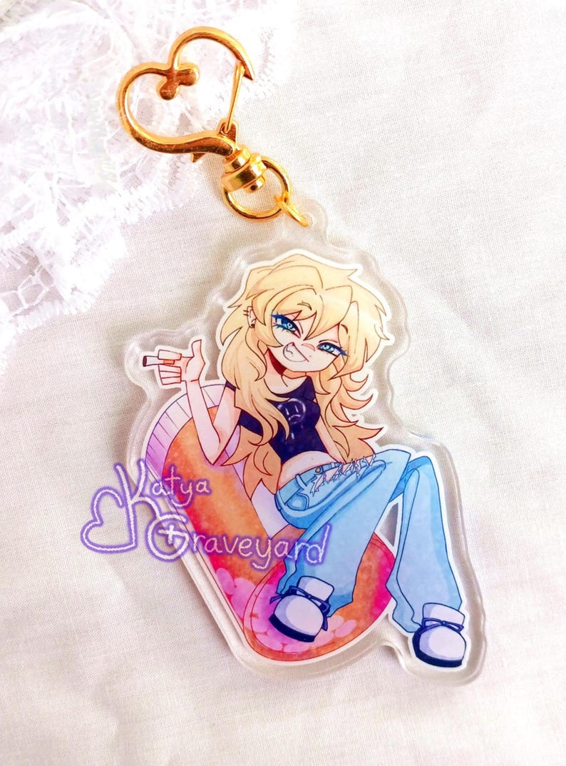 May include: Clear acrylic keyring with a cartoon illustration of a person with blonde hair, blue eyes, and blue jeans. The keyring has a gold heart-shaped clasp and the text "Katya Graveyard".
