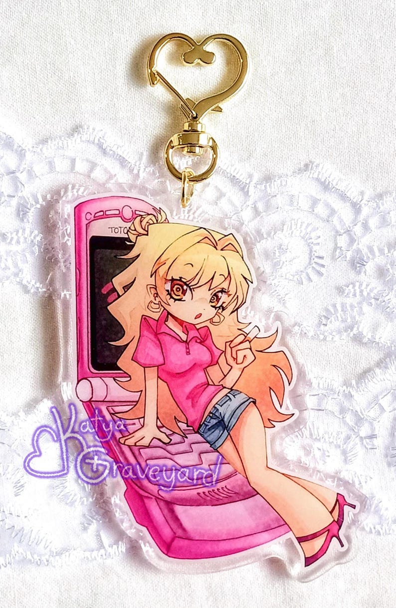 May include: A pink acrylic charm depicting an anime-style character with blonde hair, wearing a pink polo shirt and denim shorts, seated on a pink flip phone. The charm has a gold-coloured heart-shaped clasp and the text "Katya Graveyard".