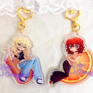 May include: Two acrylic keyrings with gold heart-shaped clasps. One shows a blonde character on a pill-shaped object, the other a red-haired character on an orange slice. Both have the text "Katya Graveyard".