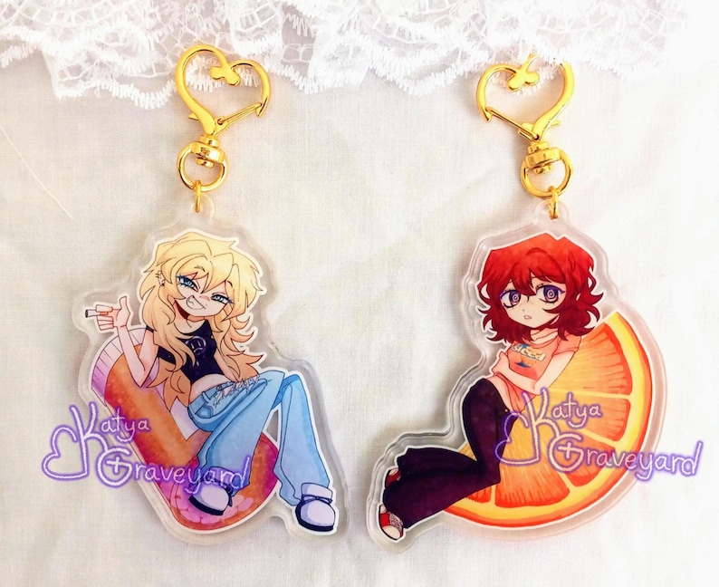 May include: Two acrylic keyrings with gold heart-shaped clasps. One shows a blonde character on a pill-shaped object, the other a red-haired character on an orange slice. Both have the text "Katya Graveyard".
