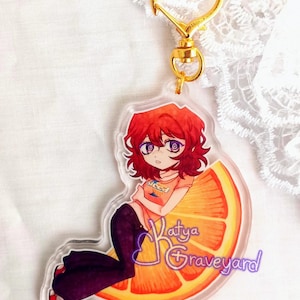 May include: A clear acrylic keychain with an anime-style character with red hair and purple eyes, sitting on an orange slice. The keychain has a gold heart-shaped clasp and a gold ring. The text "Katya Graveyard" is visible on the keychain.