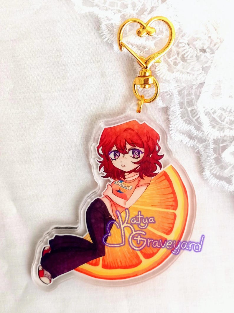 May include: A clear acrylic keychain with an anime-style character with red hair and purple eyes, sitting on an orange slice. The keychain has a gold heart-shaped clasp and a gold ring. The text "Katya Graveyard" is visible on the keychain.