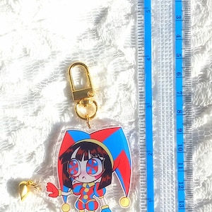 May include: A clear acrylic keychain featuring a cartoon jester character with a red, blue, and yellow color scheme. The keychain is approximately 3 inches in height and includes a gold-colored clasp and heart-shaped charm. A blue ruler is in the background.