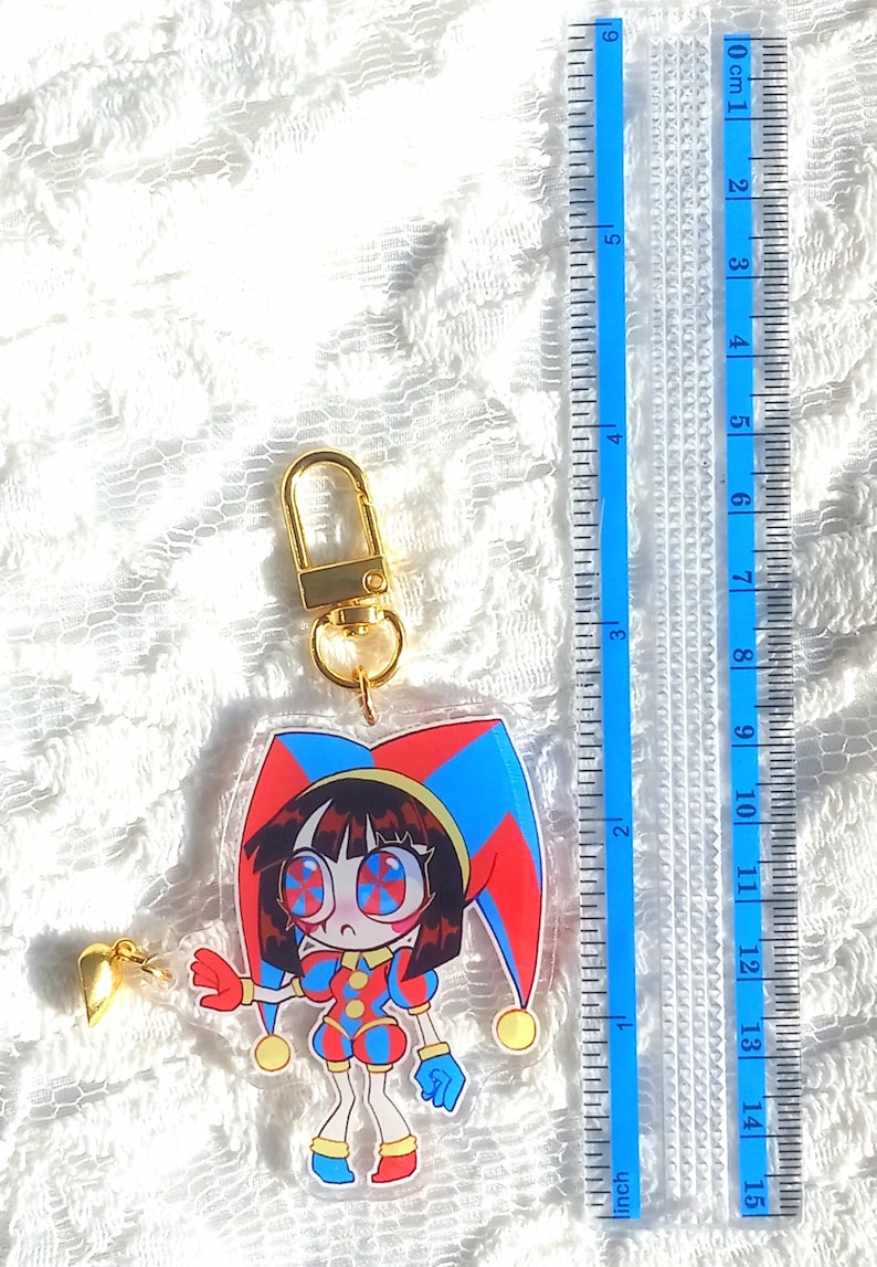 May include: A clear acrylic keychain featuring a cartoon jester character with a red, blue, and yellow color scheme. The keychain is approximately 3 inches in height and includes a gold-colored clasp and heart-shaped charm. A blue ruler is in the background.