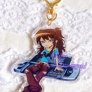 May include: A clear acrylic keychain with an anime-style character with brown hair and blue eyes, wearing a blue shirt, sitting on a flip phone. It has a gold heart-shaped clasp and the text "Latya Graveyard".