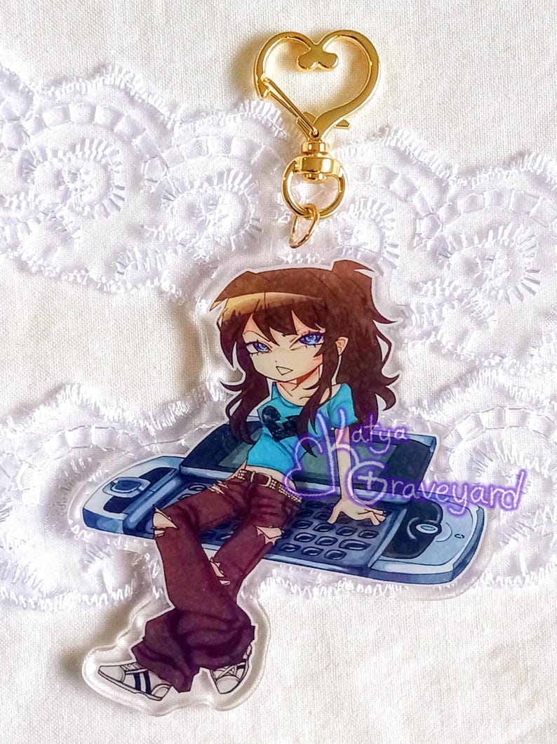 May include: A clear acrylic keychain with an anime-style character with brown hair and blue eyes, wearing a blue shirt, sitting on a flip phone. It has a gold heart-shaped clasp and the text "Latya Graveyard".
