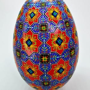 Peut inclure: Un œuf décoratif avec un motif géométrique vibrant en rouge, bleu, jaune et vert. L'œuf a une finition brillante et repose sur un support blanc. Le texte "Pysanky By Arsen Romanyuk" est en bas.