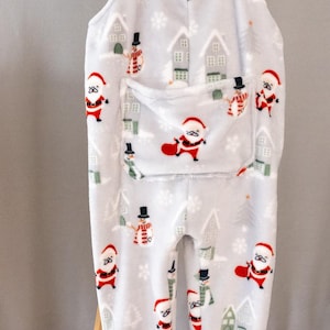Peut inclure: Combinaison de bébé gris clair avec un motif sur le thème de Noël. Le motif comprend le Père Noël, des bonhommes de neige, des maisons et des flocons de neige. La combinaison a une poche et une bordure rouge aux chevilles.