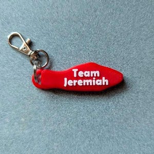 May include: A red keychain with the text "Team Jeremiah" in white. The keychain has a silver-colored clasp for attaching to keys or a bag. The background is a textured, light blue surface.