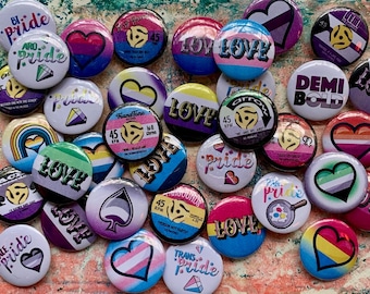 May include: A collection of colorful button pins with various pride flags, hearts, and music-themed designs. The pins feature phrases like "Bi Pride", "Aro Pride", "Love", "Pride", "Trans Pride", "Demi Bold", and "45 RPM".