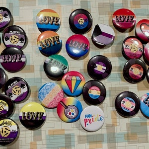 Può includere: Una collezione di spille colorate con vari disegni, tra cui cuori, giradischi e le parole "Love" e "Pride".