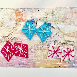 May include: A selection of handmade earrings in various colours. The earrings feature a diamond shape with a retro starburst design. Colours include light blue, hot pink, and white, with silver-tone ear wires and decorative beads. A unique accessory.