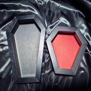 Può includere: Due scatole espositive a forma di bara con cornici in legno nero. La scatola più grande presenta una base interna grigio chiaro, mentre la più piccola presenta una base interna rosso vivace. Entrambe hanno una forma esagonale.
