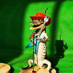May include: A taxidermied alligator wearing a red cowboy hat and holding a blue electric guitar. The alligator is standing on a wooden platform with a microphone stand.