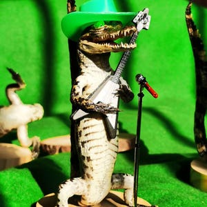 May include: A taxidermied alligator wearing a green cowboy hat and holding a toy guitar. The alligator is standing on a wooden base with a microphone stand and a red microphone.