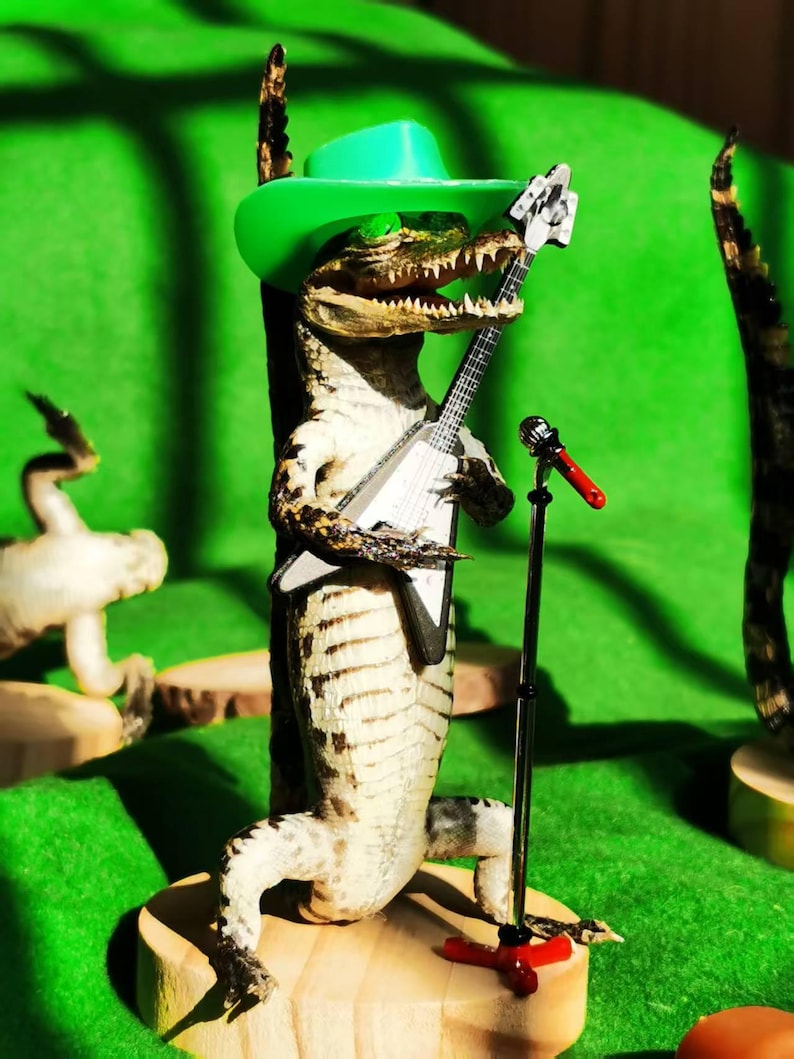 May include: A taxidermied alligator wearing a green cowboy hat and holding a toy guitar. The alligator is standing on a wooden base with a microphone stand and a red microphone.