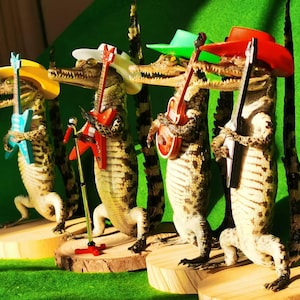 May include: Four taxidermied alligators wearing colorful hats and holding toy guitars. The alligators are arranged in a row on wooden bases.