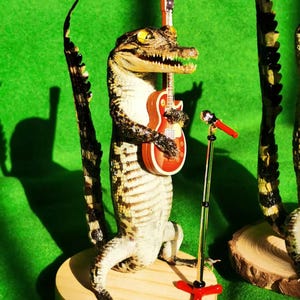 May include: A taxidermied alligator holding a miniature electric guitar and a microphone stand on a wooden base. The alligator is posed as if it is playing the guitar. The background is a green surface.
