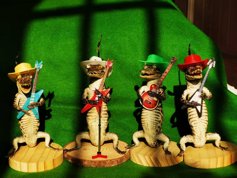 May include: Four taxidermied alligators wearing hats and playing toy guitars. The alligators are wearing a variety of hats, including a cowboy hat, a green fedora, a red cowboy hat, and a yellow cowboy hat. The guitars are all different colors and styles.
