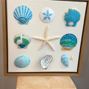 Könnte beinhalten: Ein gerahmtes Kunstwerk mit einer Sammlung von Muscheln und einem Seestern. Die Muscheln sind in Blau-, Grün- und Weißtönen mit goldenen Akzenten bemalt. Das Kunstwerk wird vor einem neutralen Hintergrund präsentiert und erzeugt ein maritimes Thema.