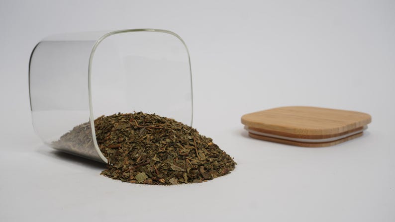 Może przedstawiać: Przezroczysty szklany słoik, przechylony na bok, wysypuje suszone zielone zioła na białą powierzchnię. W pobliżu leży kwadratowa, drewniana pokrywka. Zioła wydają się być herbatą liściastą lub podobnym składnikiem kulinarnym.