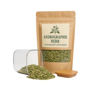 Puede incluir: Una bolsa de papel marrón con una etiqueta que dice "ANDROGRAPHIS HERB (Andrographis paniculata)". La bolsa contiene hierbas verdes secas. Un frasco de vidrio con una tapa de madera está derramando las hierbas secas sobre una superficie blanca.