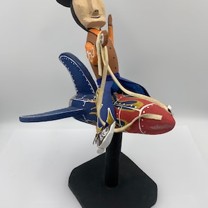 May include: A whimsical wooden sculpture of a figure wearing a black hat and riding a rocket. The rocket is painted in red and blue with gold accents. The figure has a painted face and is holding a rope.