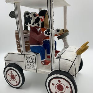 May include: A whimsical, hand-painted golf cart sculpture. The white cart features black and red wheels, a canopy, and two figures: a Dalmatian-like dog and a person. Golf clubs are visible in the back. The cart is decorated with black lines and a decorative design.