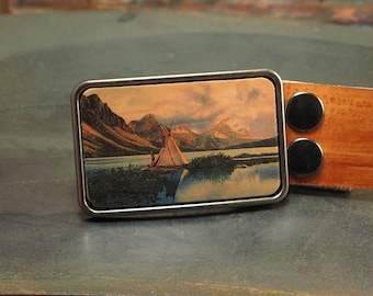 May include: A silver belt buckle with a vintage postcard image of a lake and mountains. The postcard shows a small tent on the lake with a person standing nearby. The buckle is attached to a brown leather belt.