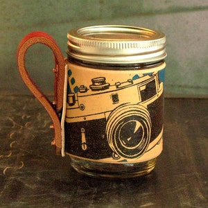 Könnte beinhalten: Ein Glas-Einmachglas mit einem Ledergriff und einem Vintage-Kamera-Design, das auf die Seite gedruckt ist. Das Glas hat einen silbernen Deckel.