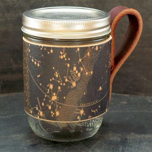 May include: A glass mason jar with a leather handle and a black and gold constellation print wrapped around it. The print features the constellation Canis Major and the text "Equinoctial".