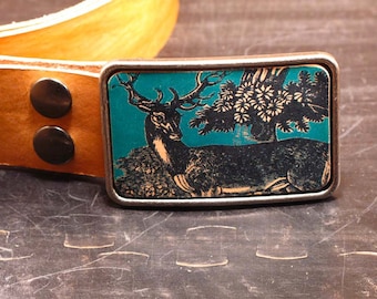 May include: A brown leather belt with a silver buckle featuring a black and white image of a deer in a forest setting. The background of the image is teal.