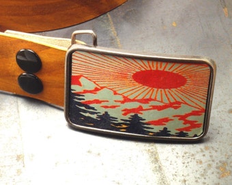 May include: A silver belt buckle featuring a colourful image of a mountain scene with a red sun rising over the peaks. The buckle has a textured, aged appearance.
