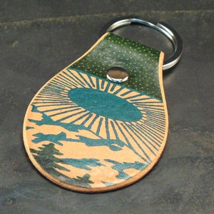 May include: A tan leather keyring with a green and blue sunburst design. The keyring has a silver metal ring.