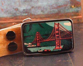May include: A silver belt buckle featuring a vintage illustration of the Golden Gate Bridge in San Francisco. The bridge is red and the background is green. The buckle is attached to a brown leather belt.