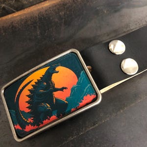 Peut inclure: Une boucle de ceinture argentée avec une image noire et verte de Godzilla debout devant un coucher de soleil. La boucle est attachée à une ceinture en cuir noir.