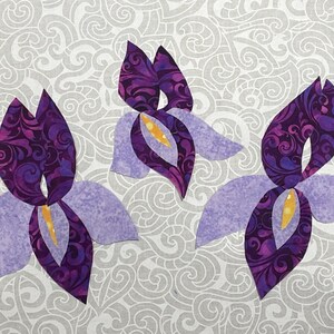 May include: Three stylized iris flower appliqu&eacute;s. The petals are a light lavender color, while the inner petals and centers are a deep purple with a swirling pattern. The centers have a yellow accent. The background is a gray and white swirling pattern.