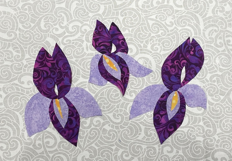 May include: Three stylized iris flower appliqu&eacute;s. The petals are a light lavender color, while the inner petals and centers are a deep purple with a swirling pattern. The centers have a yellow accent. The background is a gray and white swirling pattern.