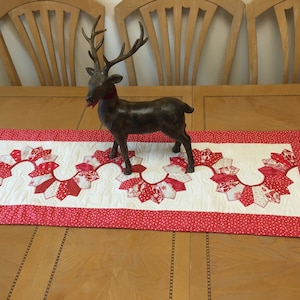 May include: A red and white quilted table runner with a scalloped edge and a white background with red polka dots. A brown reindeer figurine is placed on top of the runner.